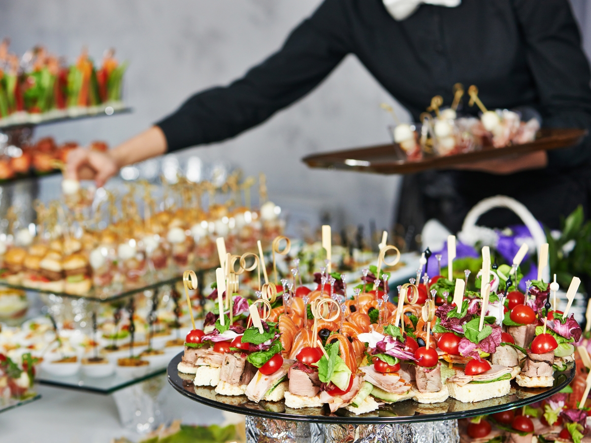 A variety of bite-sized appetizers with vegetables and meats are displayed on glass trays, perfect for corporate picnic catering, while a server in black attire places more food in the background.