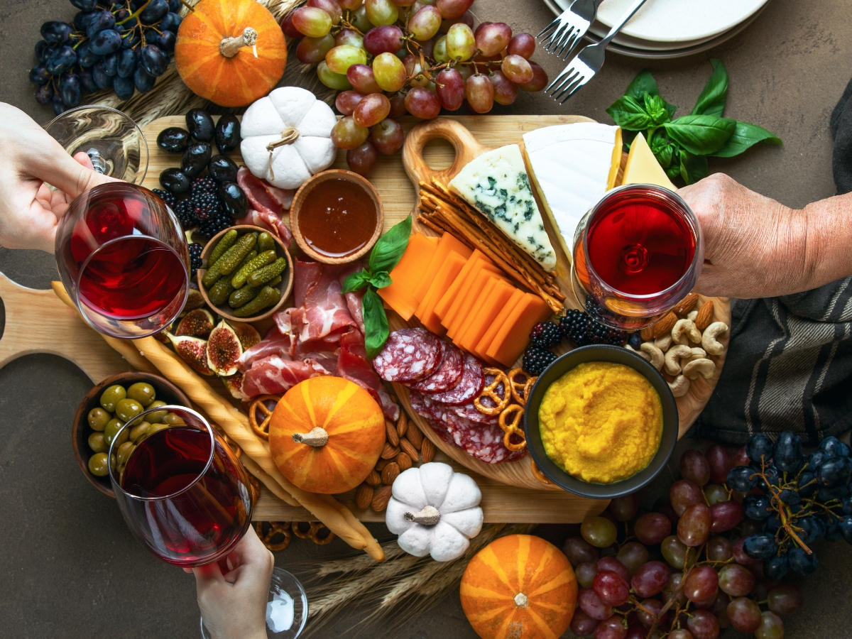 Blending Autumn Flavors Into Your Corporate Catering Menus 4 Charcuterie board with cheeses, meats, grapes, mini pumpkins, dips, nuts, and pickles—perfect for corporate catering menus. Three hands toast with red wine over the spread. Plates and utensils are set close by.