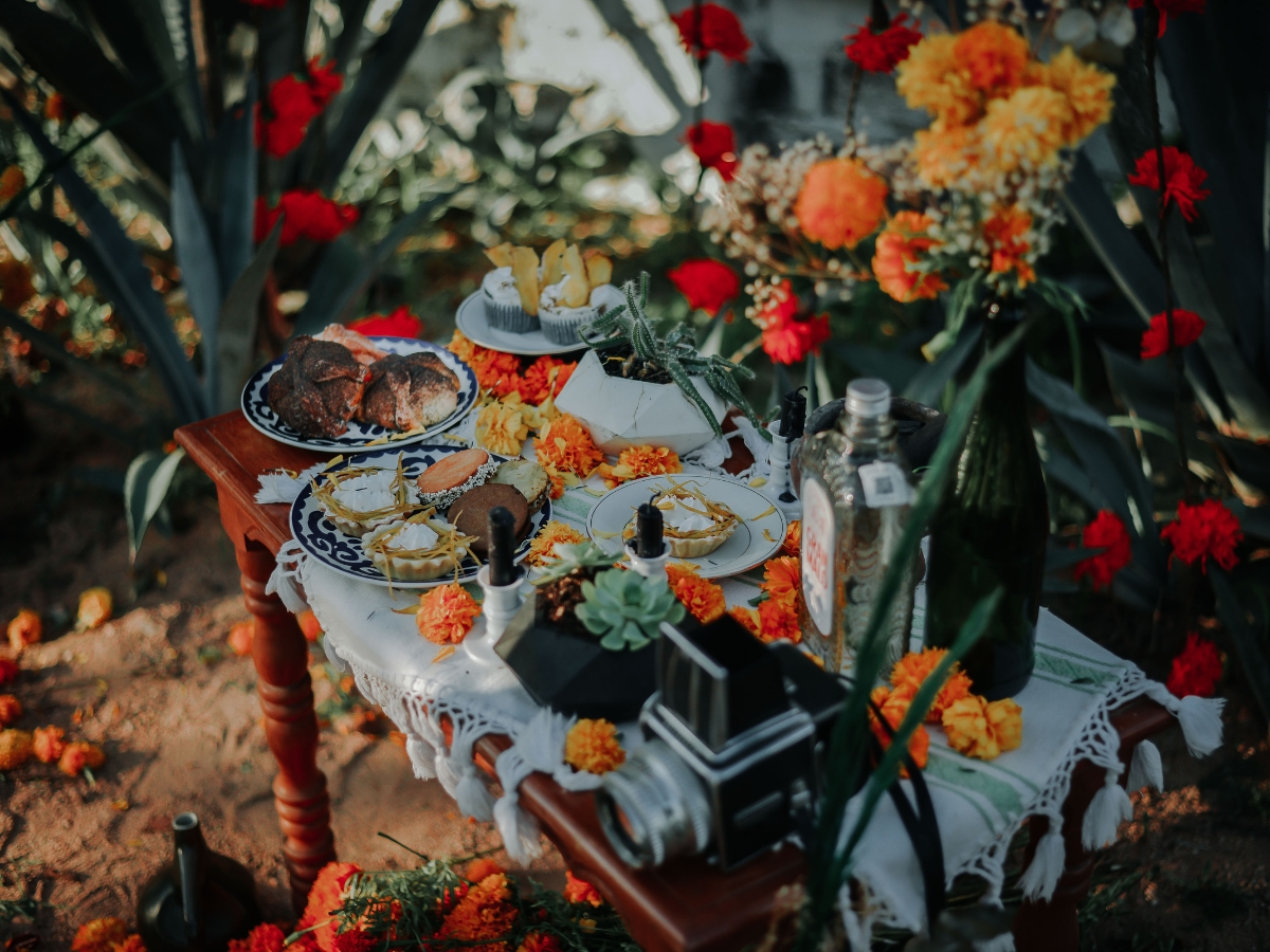 Blending Autumn Flavors Into Your Corporate Catering Menus 5 A wooden table outdoors holds plates of food, drinks, and a vintage camera, decorated with marigold flowers and surrounded by greenery—perfect inspiration for stylish corporate catering menus.