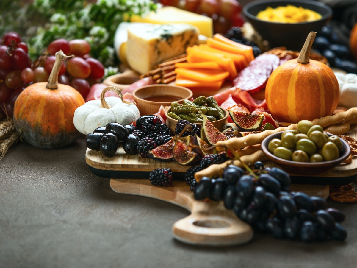 Blending Autumn Flavors Into Your Corporate Catering Menus 8 A wooden board with assorted cheeses, figs, olives, grapes, blackberries, sliced meats, pickles, crackers, and small pumpkins arranged on a table—perfect for elegant gatherings or featured on upscale corporate catering menus.