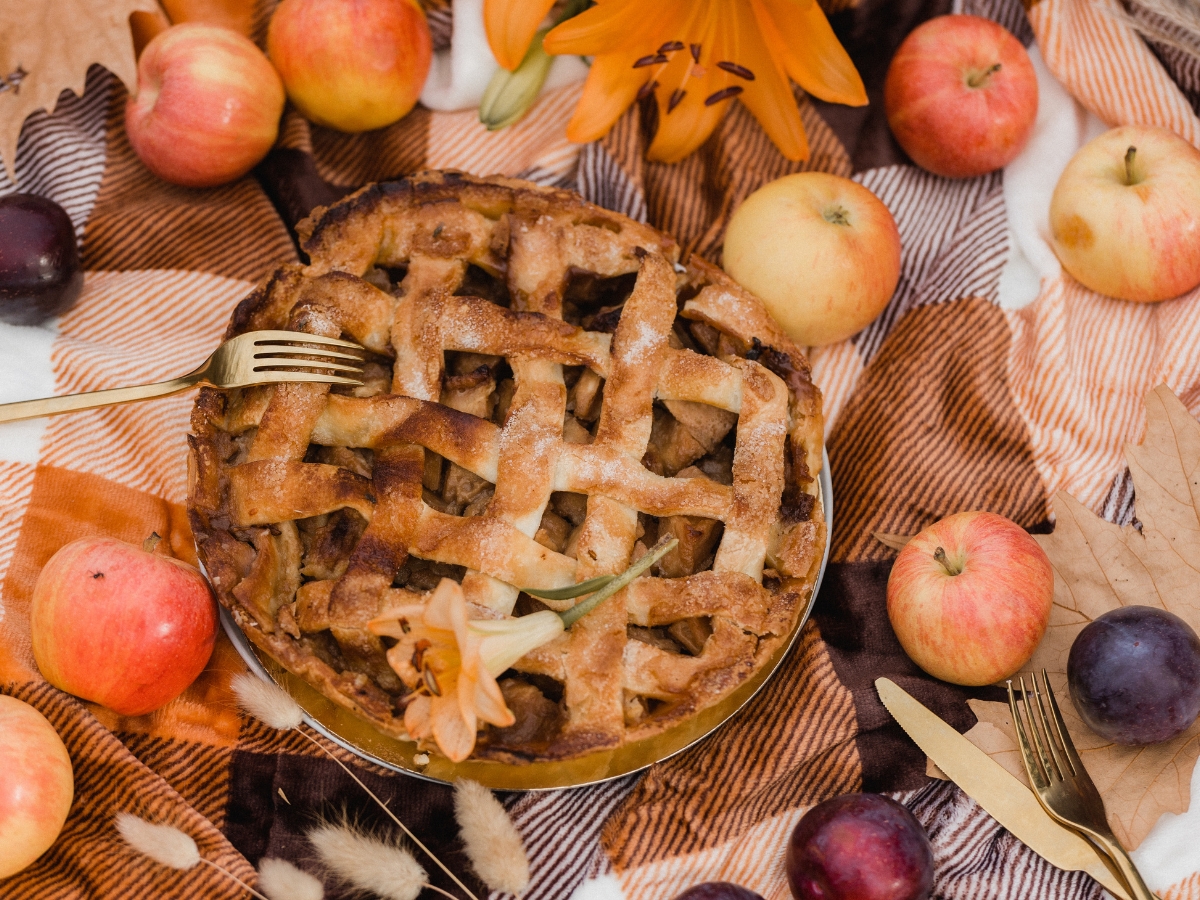 A lattice-topped pie on a checkered blanket, surrounded by apples, plums, autumn leaves, and a gold fork—perfectly styled for your fall catering menu—with an orange flower as decoration.