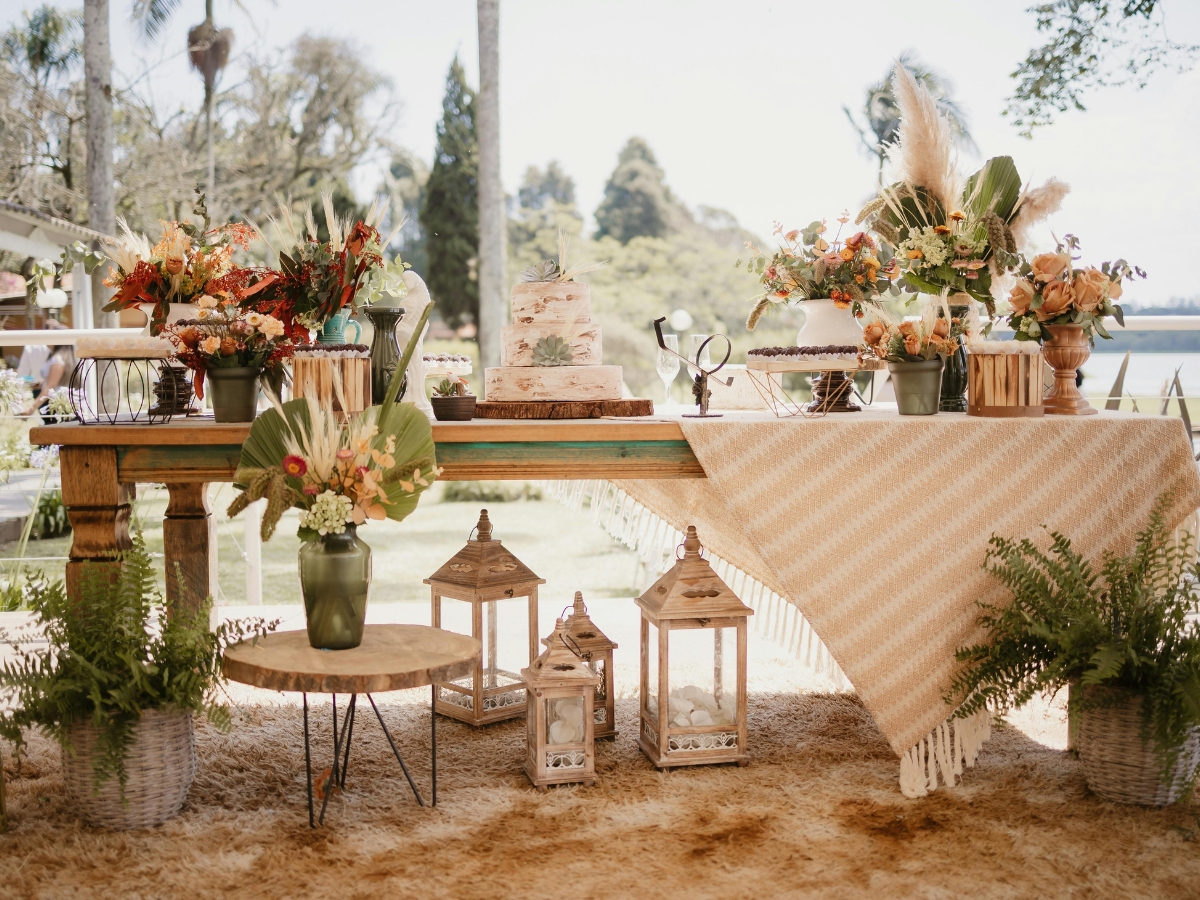 A decorated wooden table outdoors with a cake, floral arrangements, lanterns, and a beige patterned tablecloth showcases the season’s beauty—perfect for enjoying a fall catering menu amid greenery and natural light.