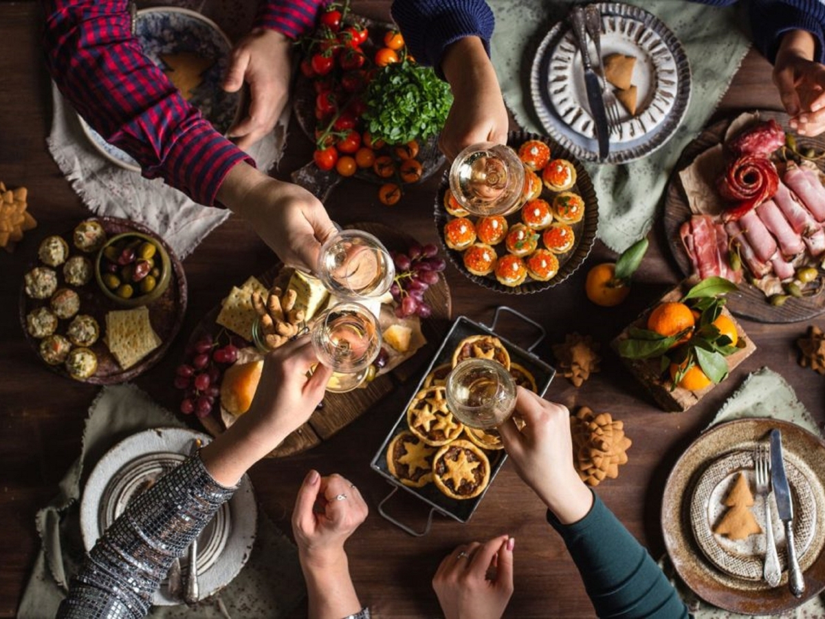 Five people clink glasses over a table set with assorted appetizers, fruit, pastries, charcuterie, and plates—a festive gathering that showcases a delicious fall catering menu.