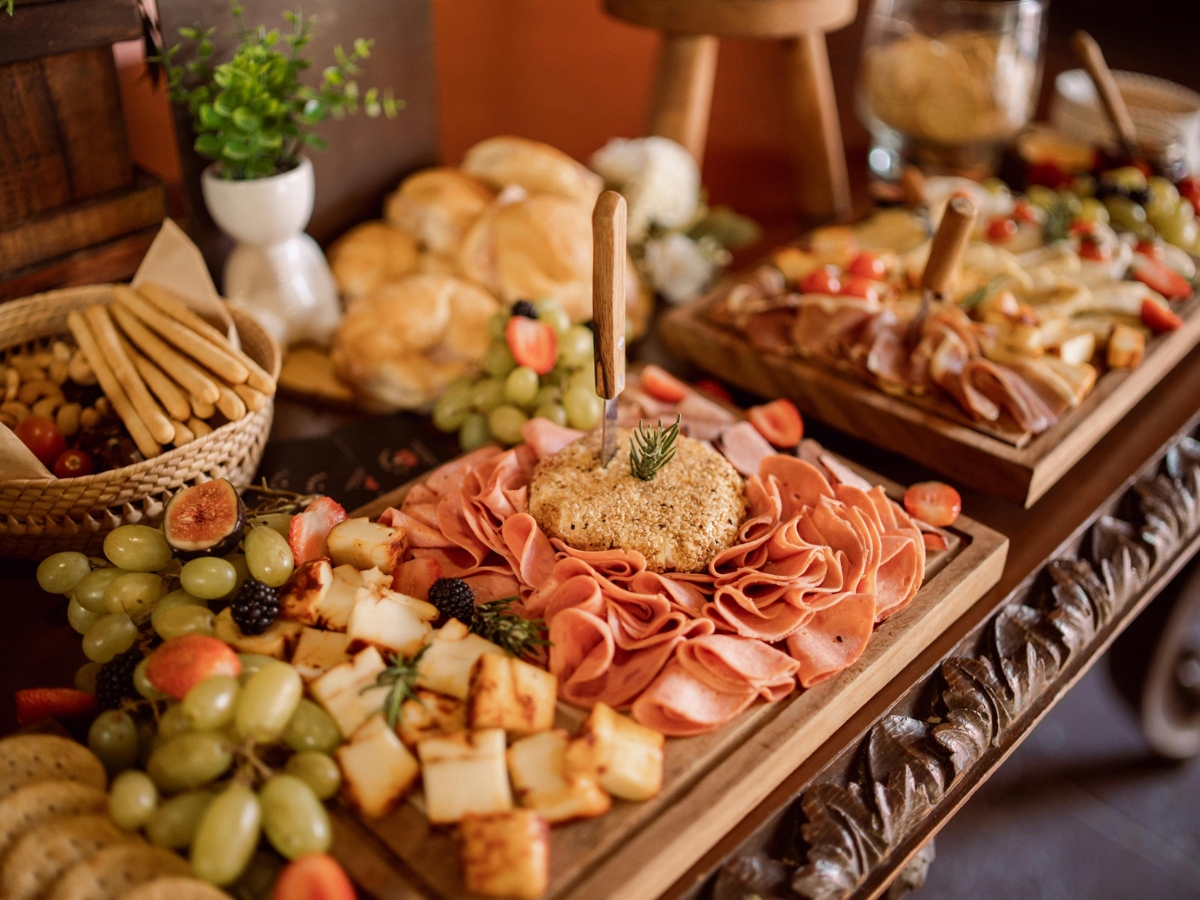 A wooden platter with sliced deli meats, cheese, grapes, figs, and bread is displayed on a decorative table, showcasing a delicious fall catering menu with additional trays of assorted foods in the background.