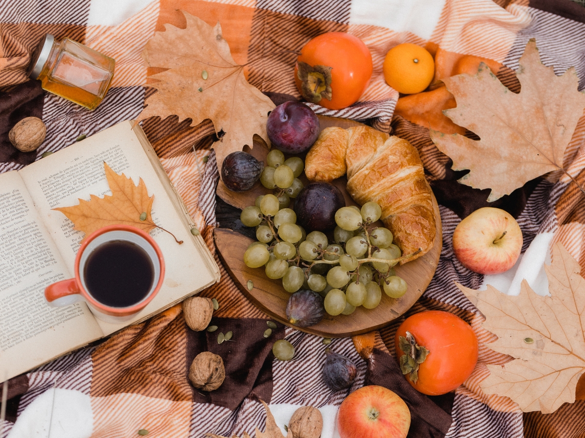 A wooden plate with grapes, figs, and a croissant sits on a plaid blanket, surrounded by apples, persimmons, walnuts, autumn leaves, coffee, a book, and a small jar—perfect inspiration for your fall catering menu.