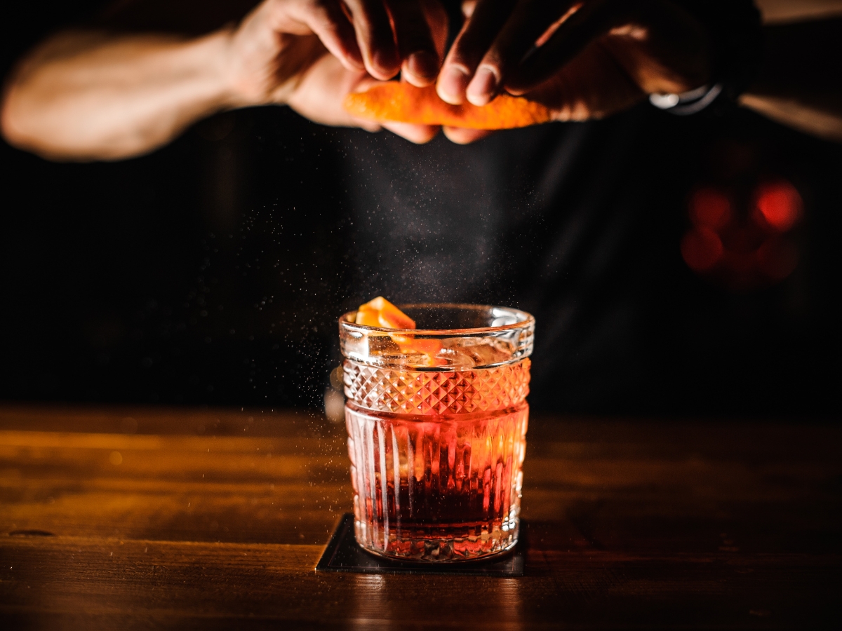 Planning a Fall-Themed Wedding Reception Menu: Seasonal Delights for Your Big Day 5 A hand squeezes an orange peel over a glass of cocktail on a wooden bar, releasing droplets onto the drink—perfect for a cozy Fall-Themed Wedding Reception.