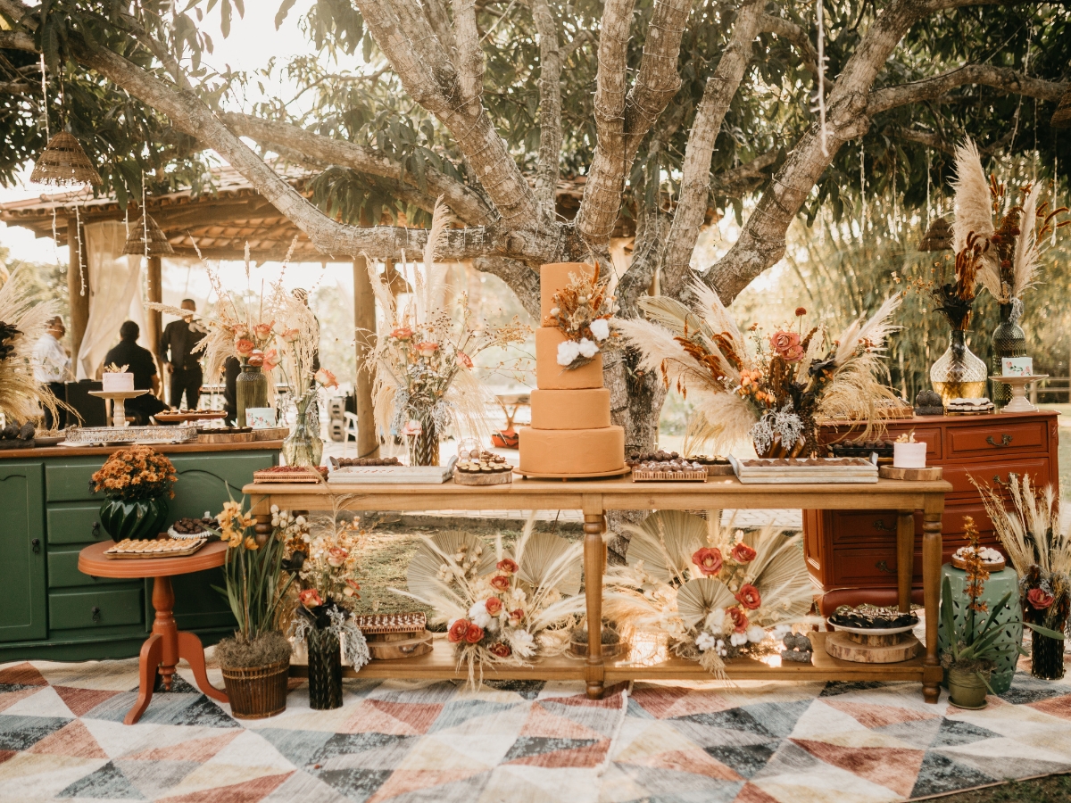 Planning a Fall-Themed Wedding Reception Menu: Seasonal Delights for Your Big Day 1 A rustic outdoor dessert table at a Fall-Themed Wedding Reception features a large tiered cake, floral arrangements, and assorted pastries beneath a tree with sunlight filtering through the branches.