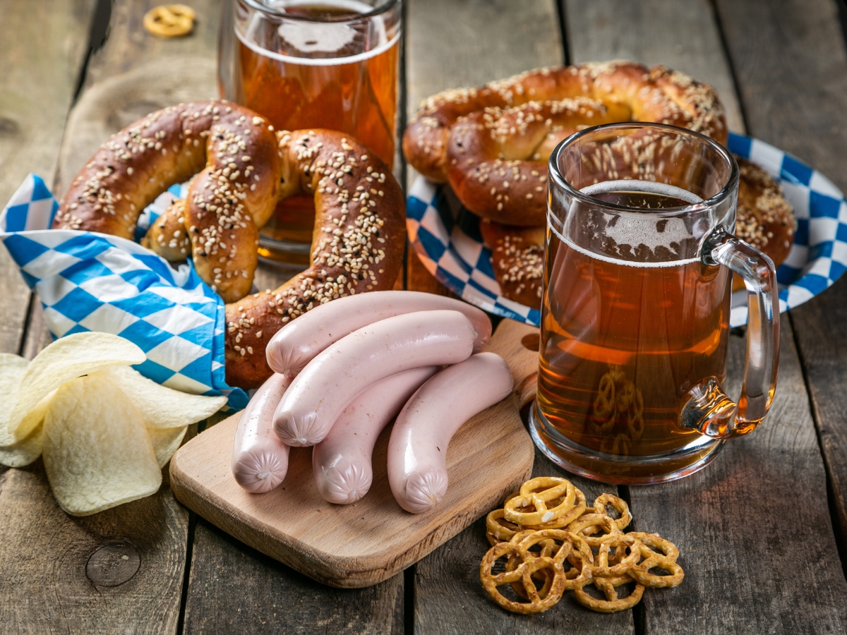 Oktoberfest Catering: Menu Ideas for a Festive Celebration 5 Two mugs of beer, soft pretzels with salt, white sausages on a wooden board, potato chips, and small pretzels create the perfect Oktoberfest Catering spread on a rustic wooden table.