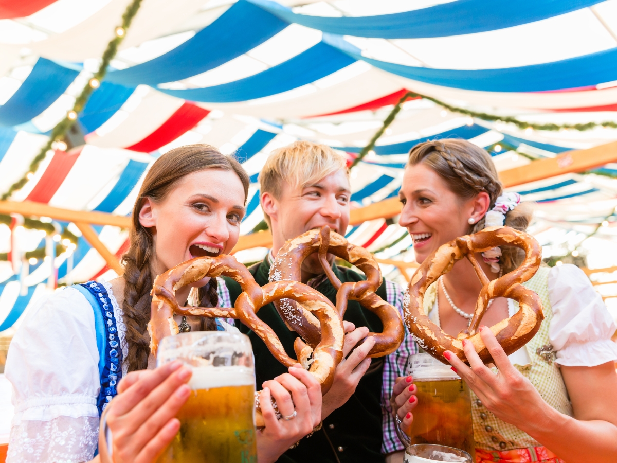 Oktoberfest Catering: Menu Ideas for a Festive Celebration 3 Three people in traditional Bavarian attire hold large pretzels and beer steins, smiling under a tent decorated with blue and white banners—perfect for an Oktoberfest Catering celebration.