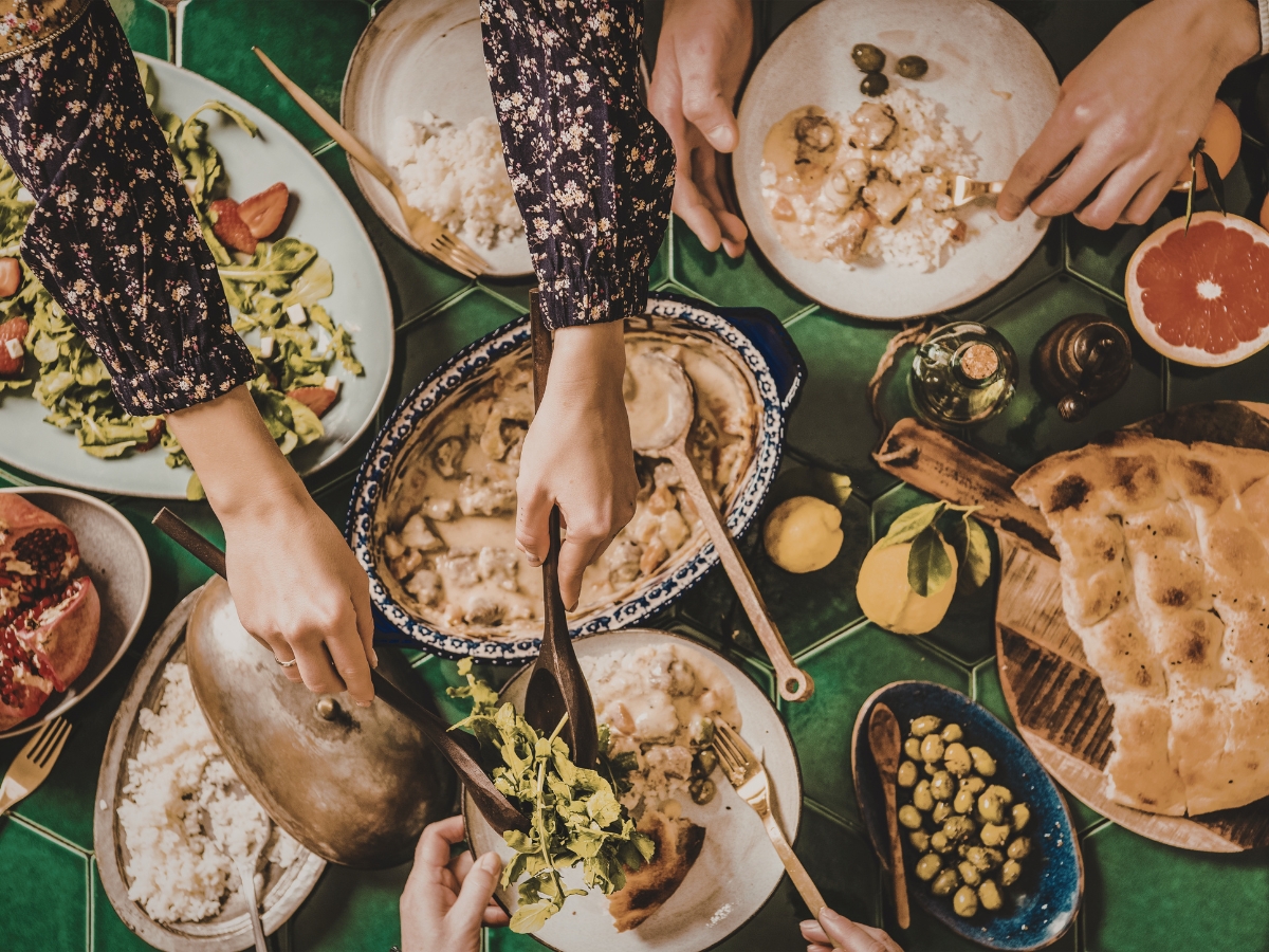 Unique Thanksgiving Catering Menu Ideas to Impress Your Guests 3 People serve themselves food from dishes including rice, salad, bread, olives, and a creamy main dish on a green-tiled table, perfect for a Thanksgiving Catering Menu.