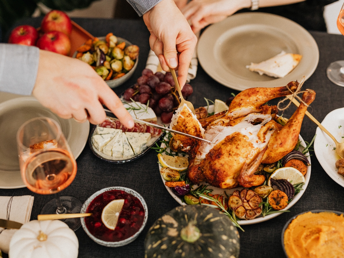 Unique Thanksgiving Catering Menu Ideas to Impress Your Guests 4 Person carving a roast chicken at a dinner table set with various side dishes, fruits, wine, and plates from the Thanksgiving Catering Menu.