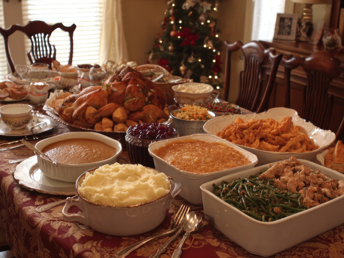 Unique Thanksgiving Catering Menu Ideas to Impress Your Guests 1 A dining table is set with various dishes from a Thanksgiving Catering Menu, including mashed potatoes, green beans, fried chicken, bread rolls, and a Christmas tree in the background.