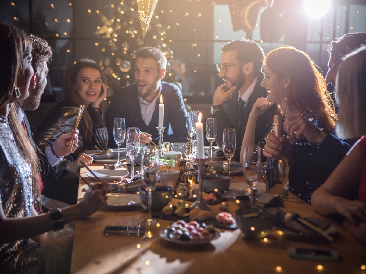 Christmas Party Catering Ideas for Intimate Gatherings 2 A group of people in formal attire sit around a decorated dinner table, talking and smiling, with a lit Christmas tree in the background—perfect inspiration for your Christmas party catering ideas.