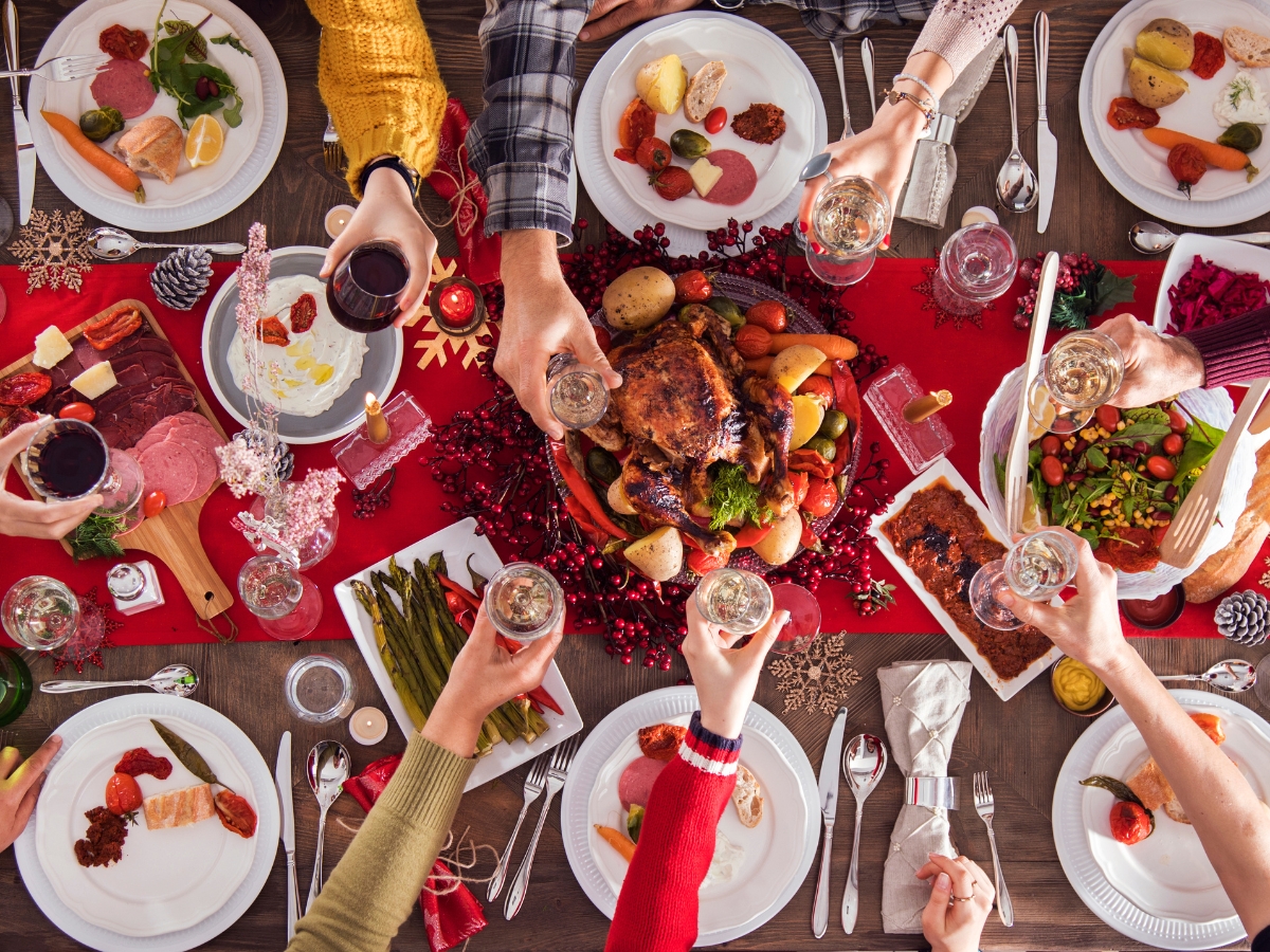 Christmas Party Catering Ideas for Intimate Gatherings 5 People seated around a table enjoy a festive meal with roast poultry, vegetables, and delicious side dishes, raising glasses for a toast—perfect inspiration for Christmas Party Catering Ideas.