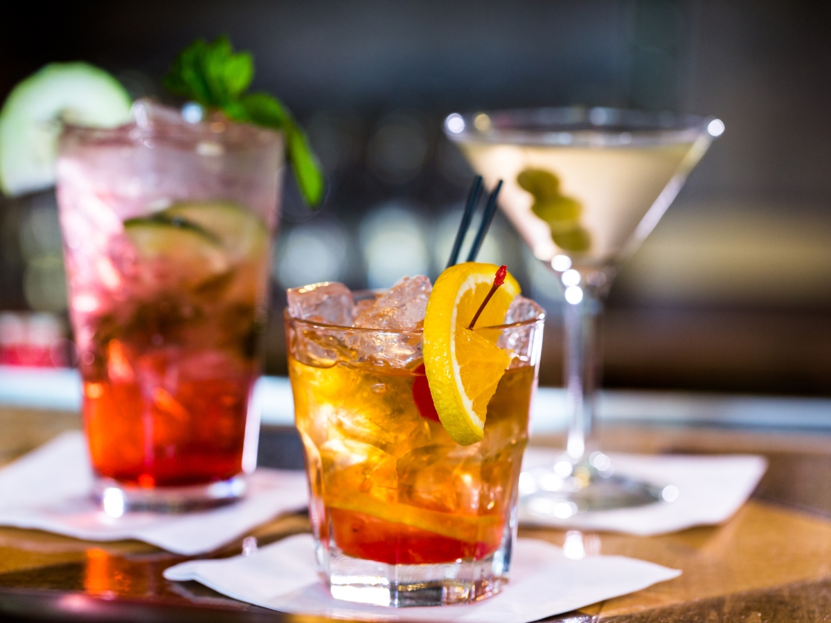 Planning a Holiday Cocktail Party: Catering Tips and Menu Ideas 3 Three cocktails on a bar at a Holiday Cocktail Party, each on a white napkin—one with lemon and cherry garnish, one with mint and cucumber, and one martini with olives.