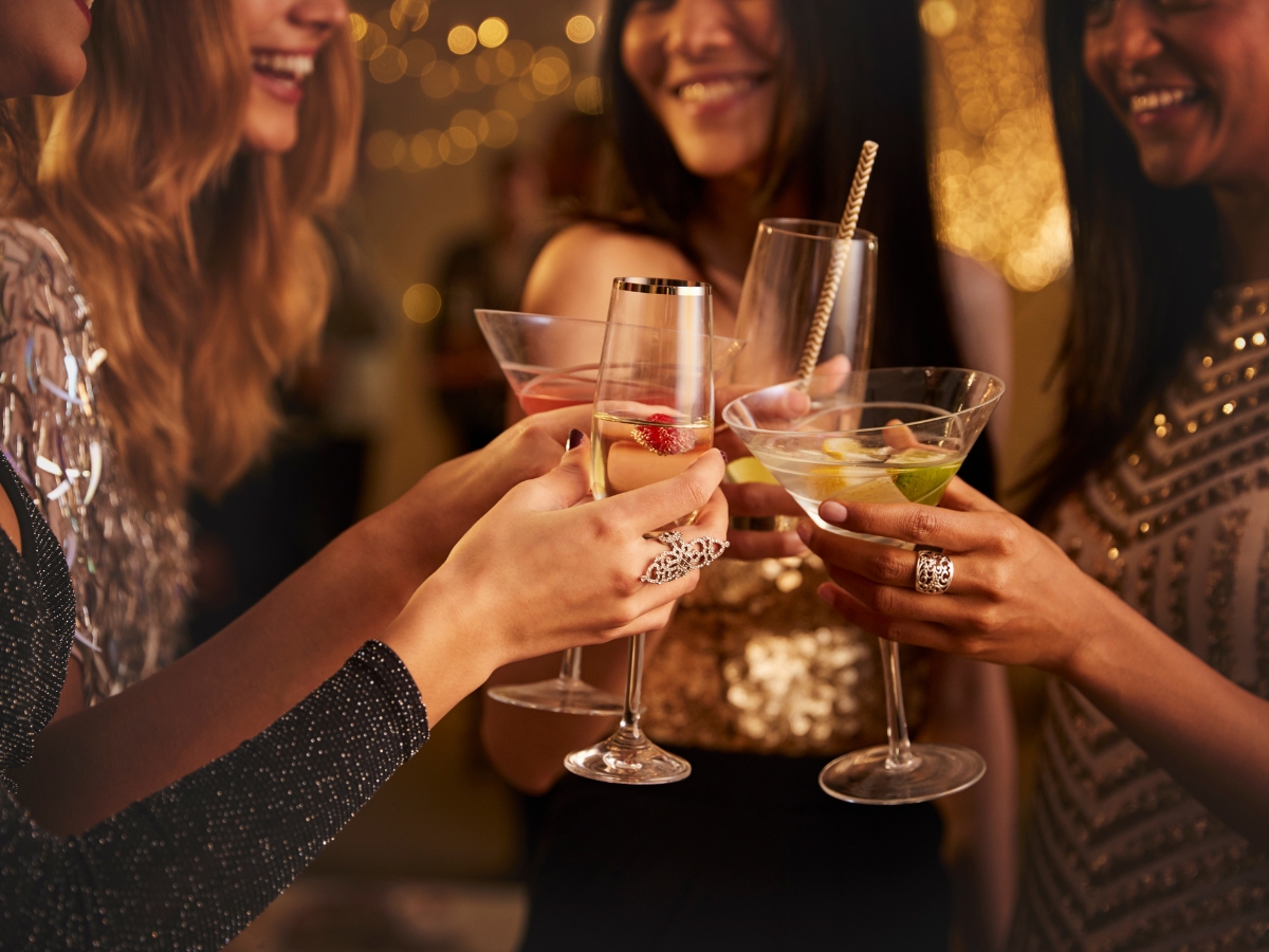 Planning a Holiday Cocktail Party: Catering Tips and Menu Ideas 1 Four women toast with various cocktails at a Holiday Cocktail Party, dressed in elegant evening wear, with warm, festive lighting glowing in the background.