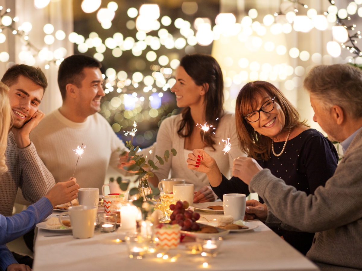 New Year's Eve Catering Tips: Create a Memorable Celebration 4 Five people sit around a table with food, drinks, and candles from a New Year’s Eve catering service, holding sparklers, smiling and talking, with blurred lights in the background.