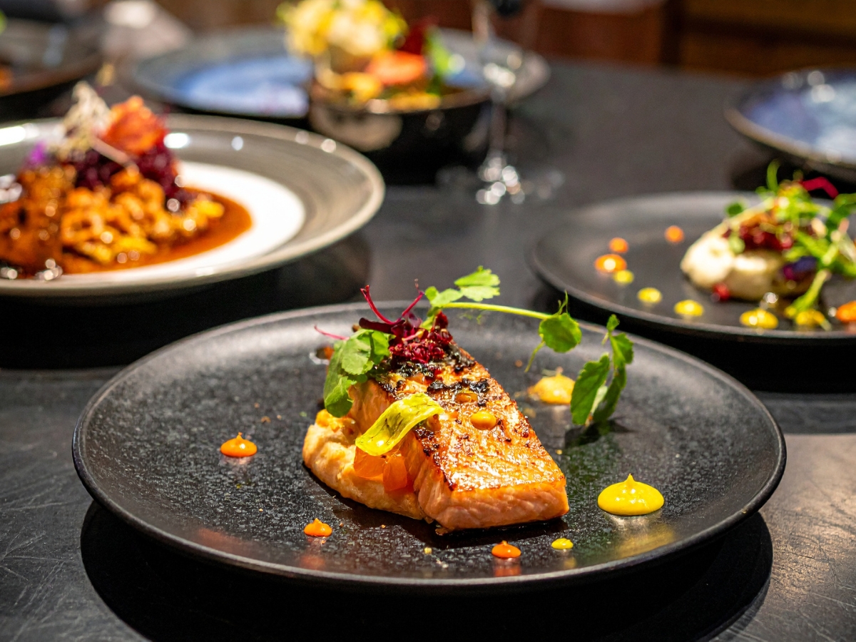 Elevating Food Presentation for Events 5 A piece of grilled salmon garnished with greens and colorful sauces showcases exquisite food presentation on a black dish, with other assorted dishes blurred in the background.
