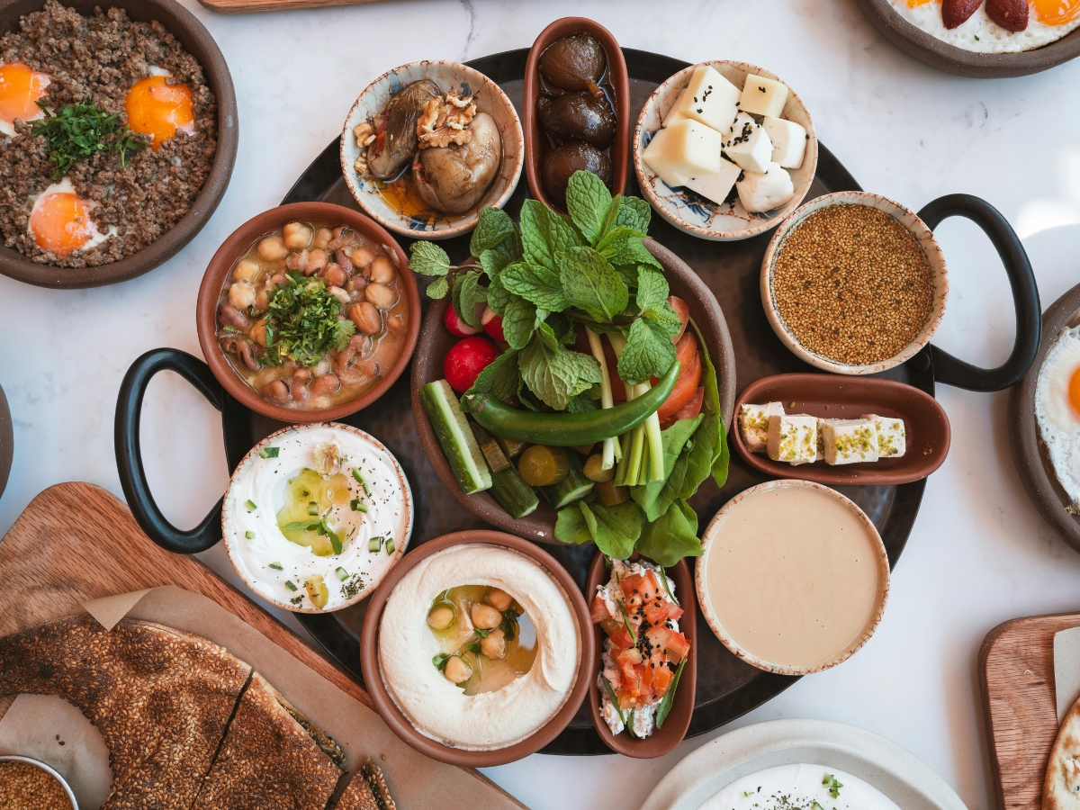 A platter with various Middle Eastern dishes, including hummus, vegetables, labneh, falafel, chickpeas, cheeses, and sauces—perfect for plant based catering—arranged on a table with other plates of food.