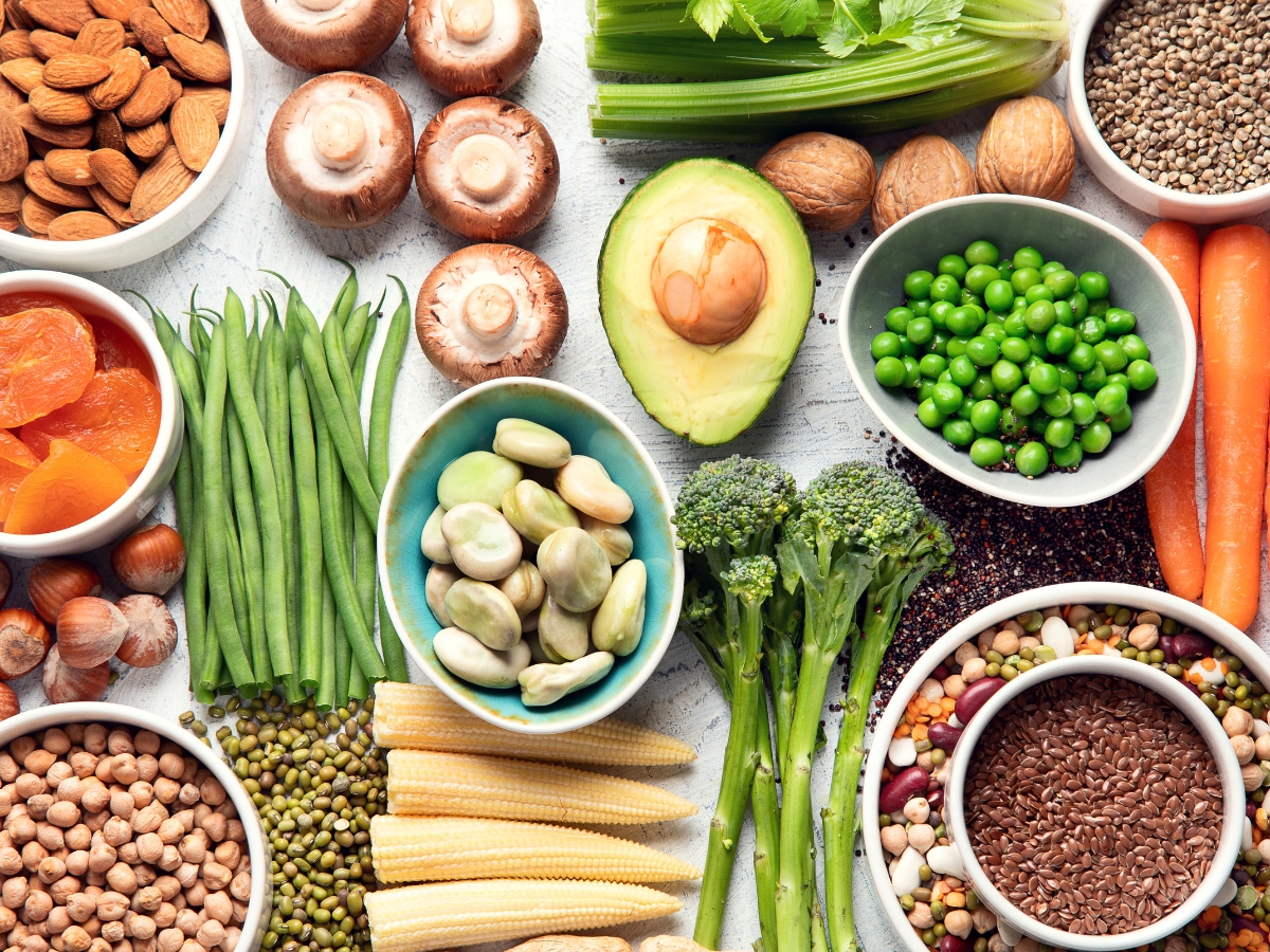 Assorted fresh vegetables, legumes, nuts, and seeds arranged in bowls and on a white surface—perfect for plant based catering—include broccoli, avocado, carrots, mushrooms, beans, peas, and almonds.