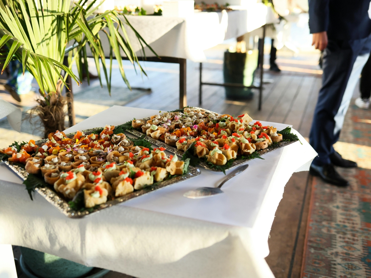 Two trays of assorted bite-sized, plant-based appetizers are arranged on a white tablecloth at an indoor event, with people and tables visible in the background—perfect for plant based catering occasions.