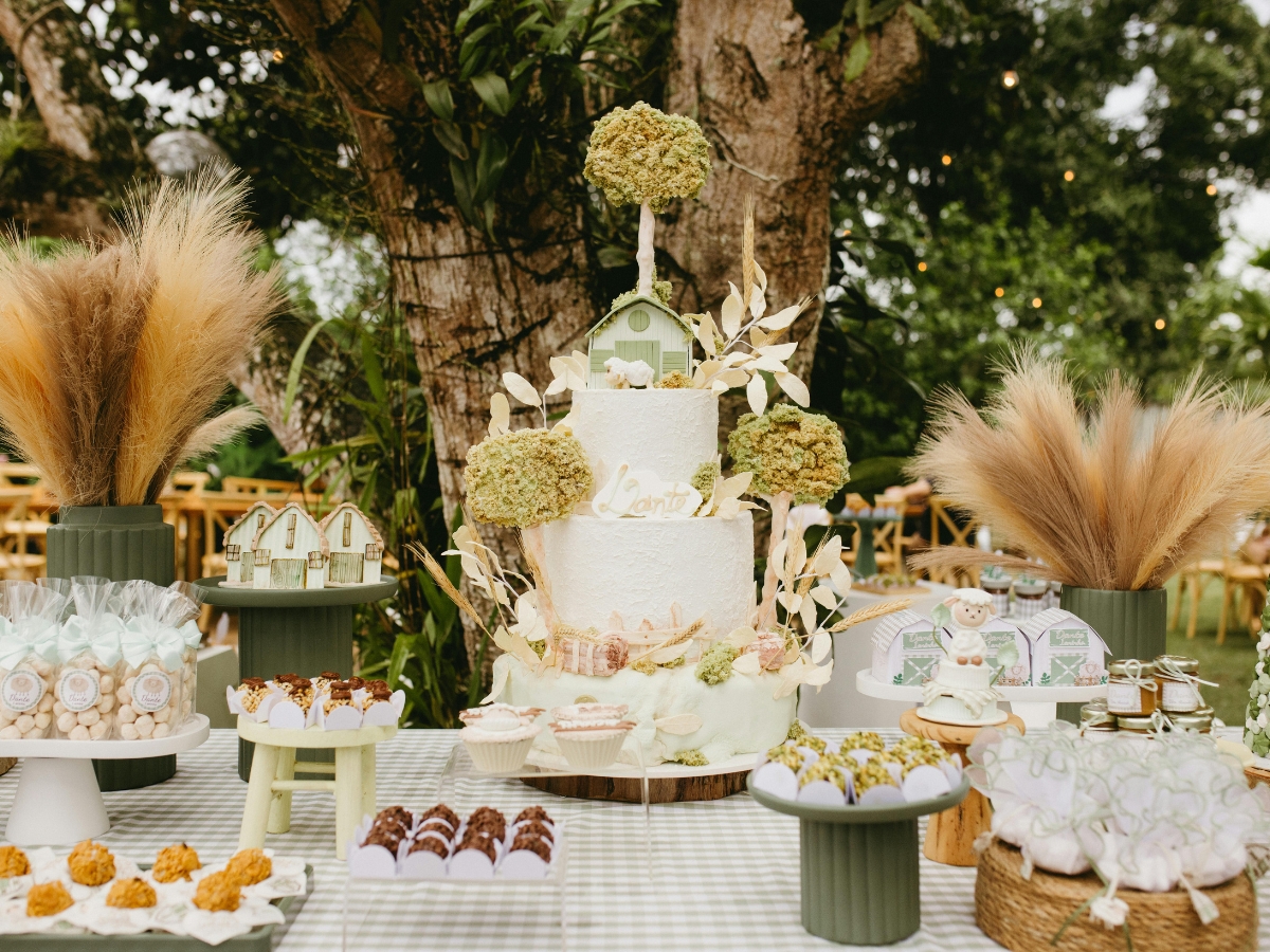 A decorated table outdoors showcases a large white cake with green accents, assorted pastries, wrapped treats, and pampas grass arrangements under a tree—perfect for elegant birthday party catering.
