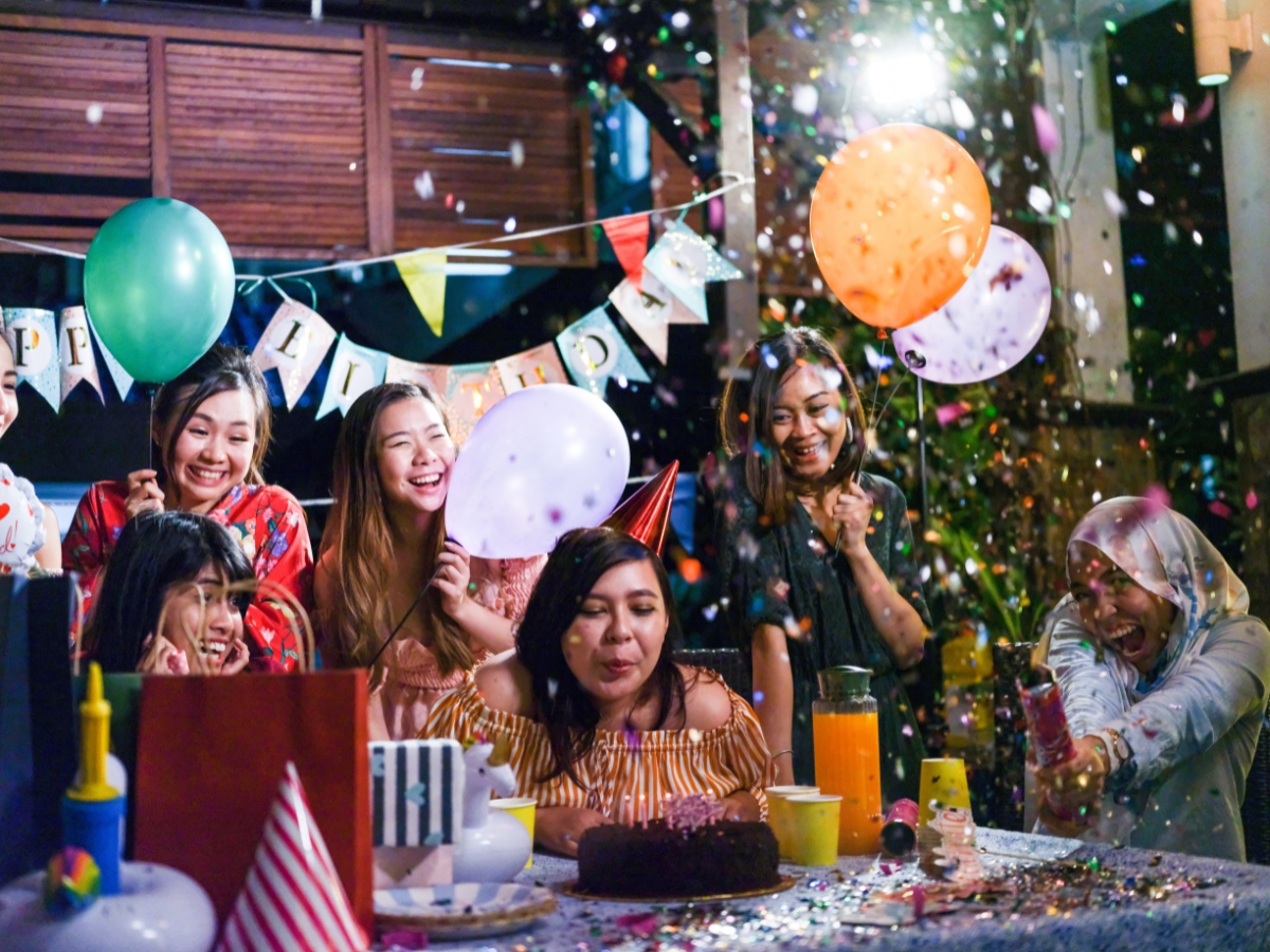 A woman wearing a party hat blows out candles on a birthday cake while friends smile, hold balloons, and confetti falls around them—perfect moments made even better with birthday party catering.