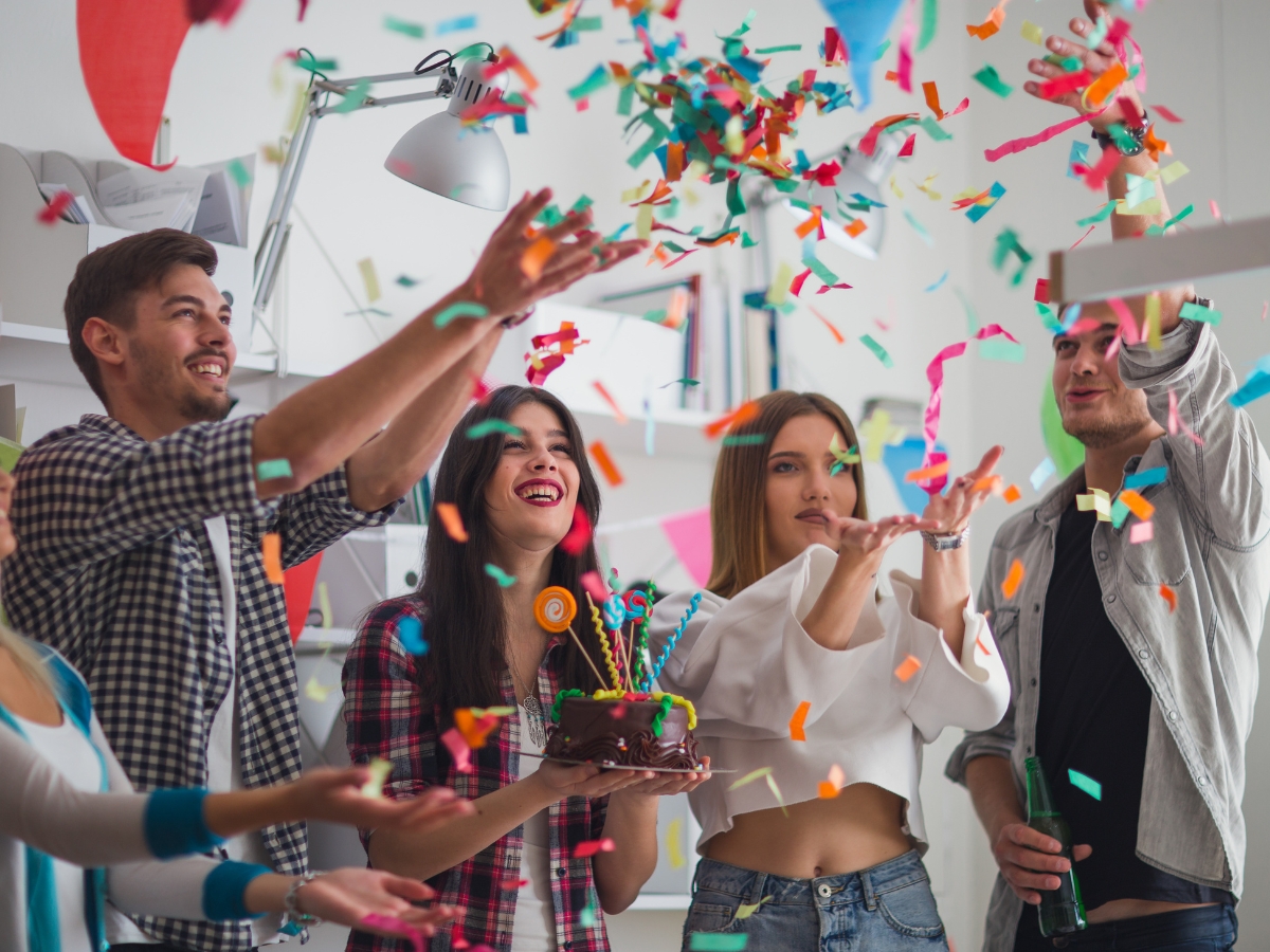 Four people celebrate indoors with colorful confetti, enjoying a vibrant Birthday Party Catering experience. One person holds a chocolate cake with lit candles as everyone appears cheerful and engaged in the moment.