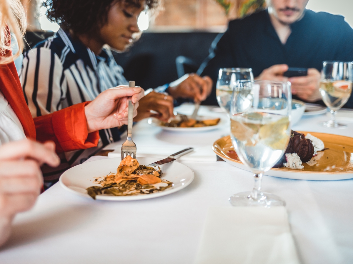 Three people sit at a table enjoying a corporate business lunch, their hands holding utensils and plates filled with greens and dessert, drinks close by.