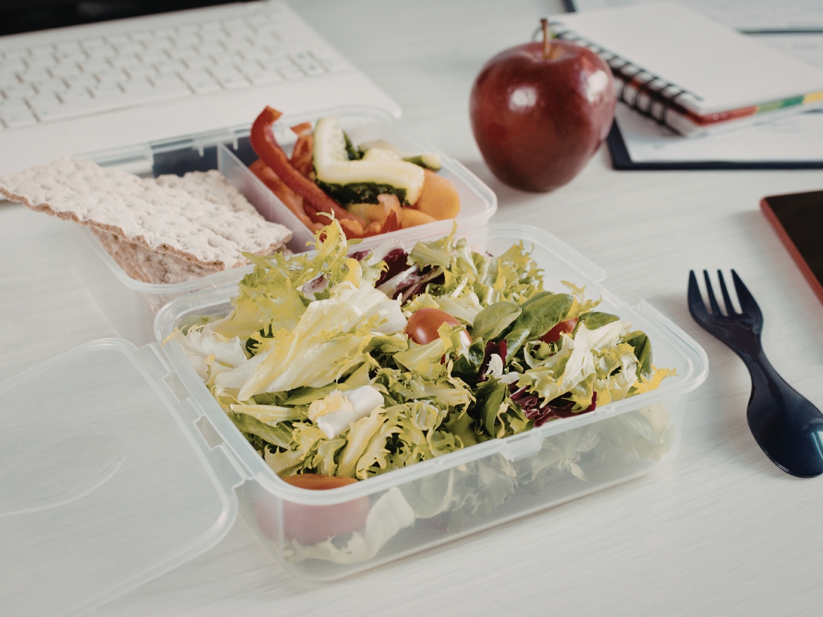 A plastic container with salad, sliced vegetables, crispbread, a red apple, and black plastic cutlery sits beside a keyboard and notebooks—perfect for a quick corporate business lunch at your desk.