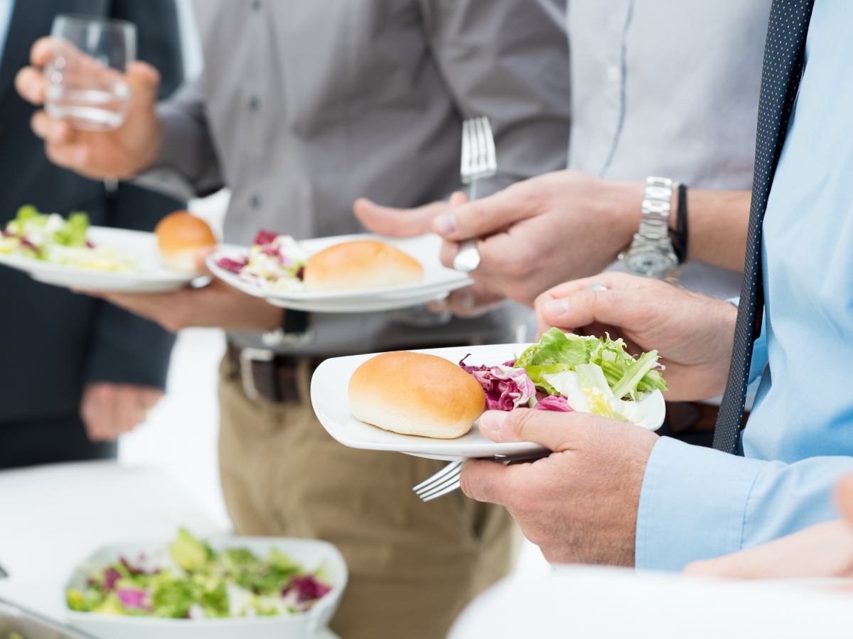 People attending a Corporate Business Lunch hold plates with salad and bread rolls at a buffet or catered event. Only their torsos and hands are visible.