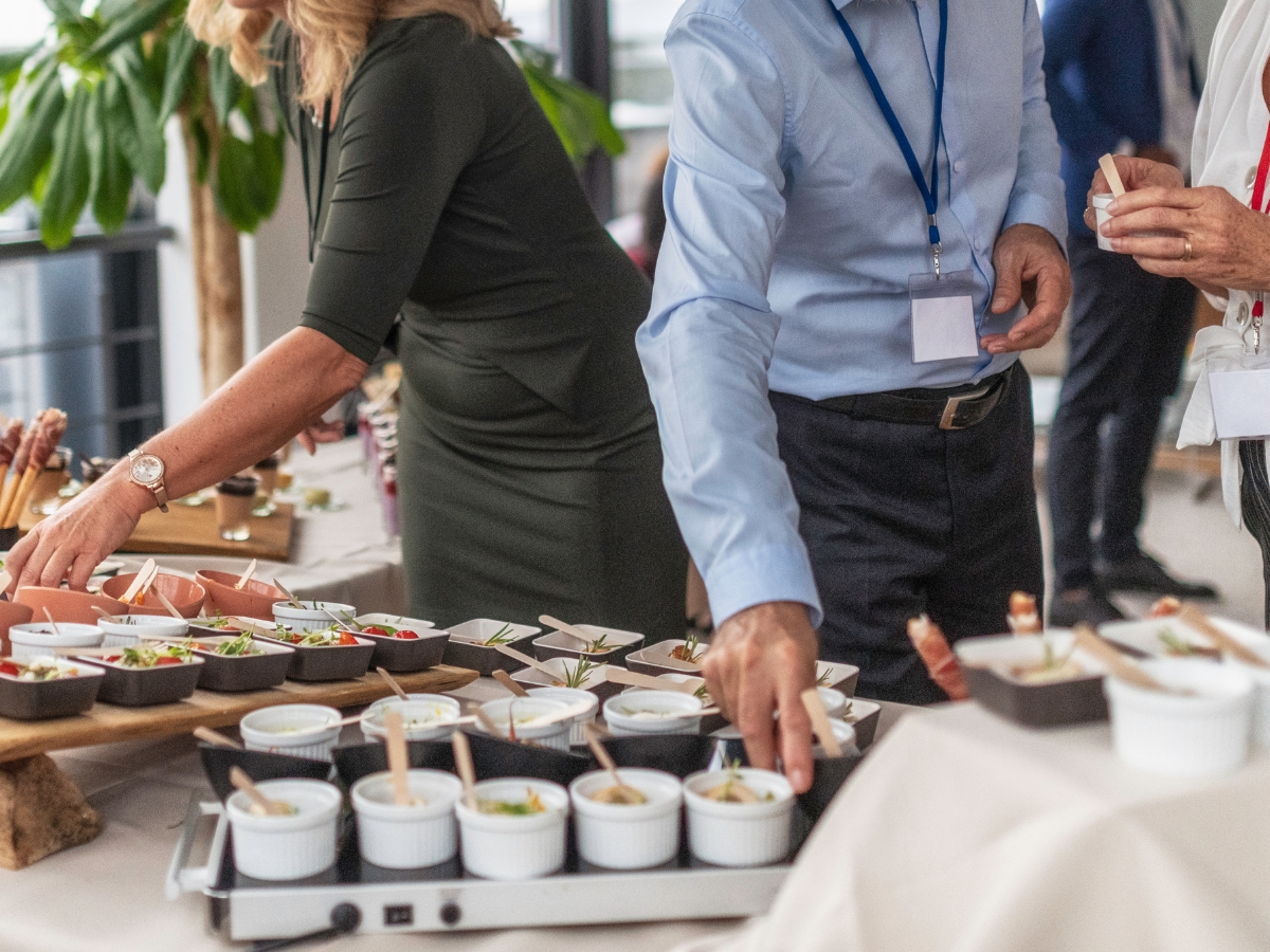 People are selecting food from a buffet table with small dishes and appetizers, each with a spoon, during what appears to be a corporate business lunch or conference.