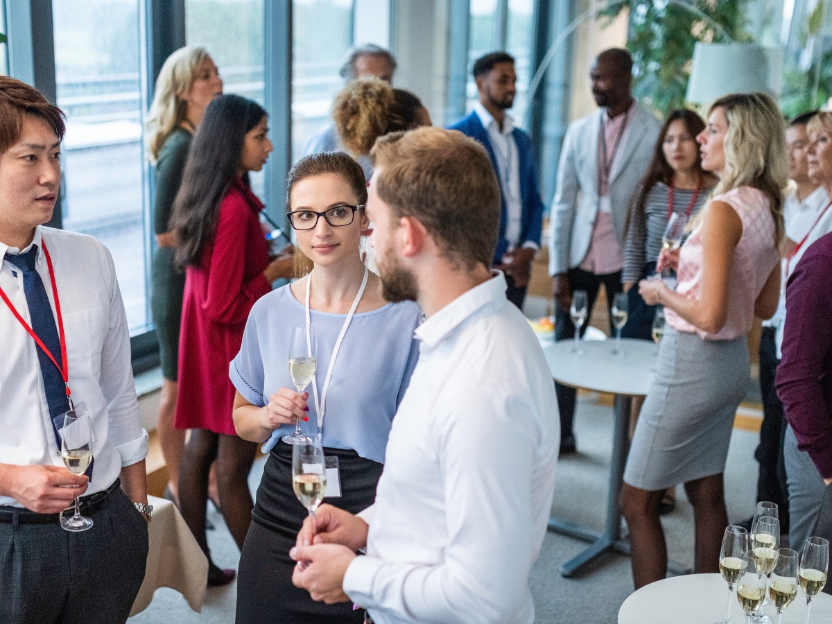 A group of professionally dressed people stand and talk while holding drinks at a corporate team building networking event in a modern, bright room.