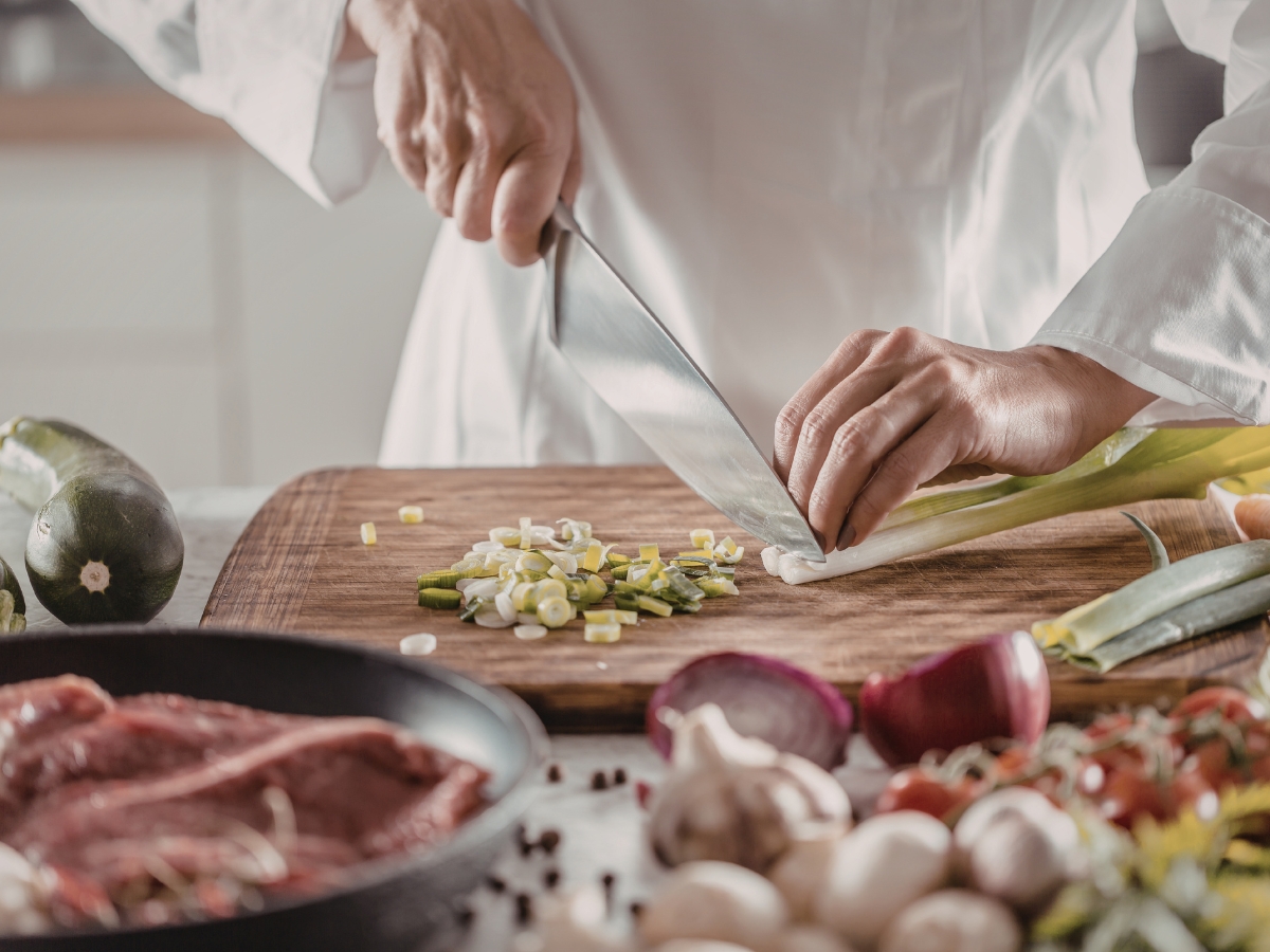 Person in a white chef's coat chopping green onions on a wooden cutting board, surrounded by various vegetables and raw meat, skillfully preparing ingredients for different cooking methods in a kitchen setting.