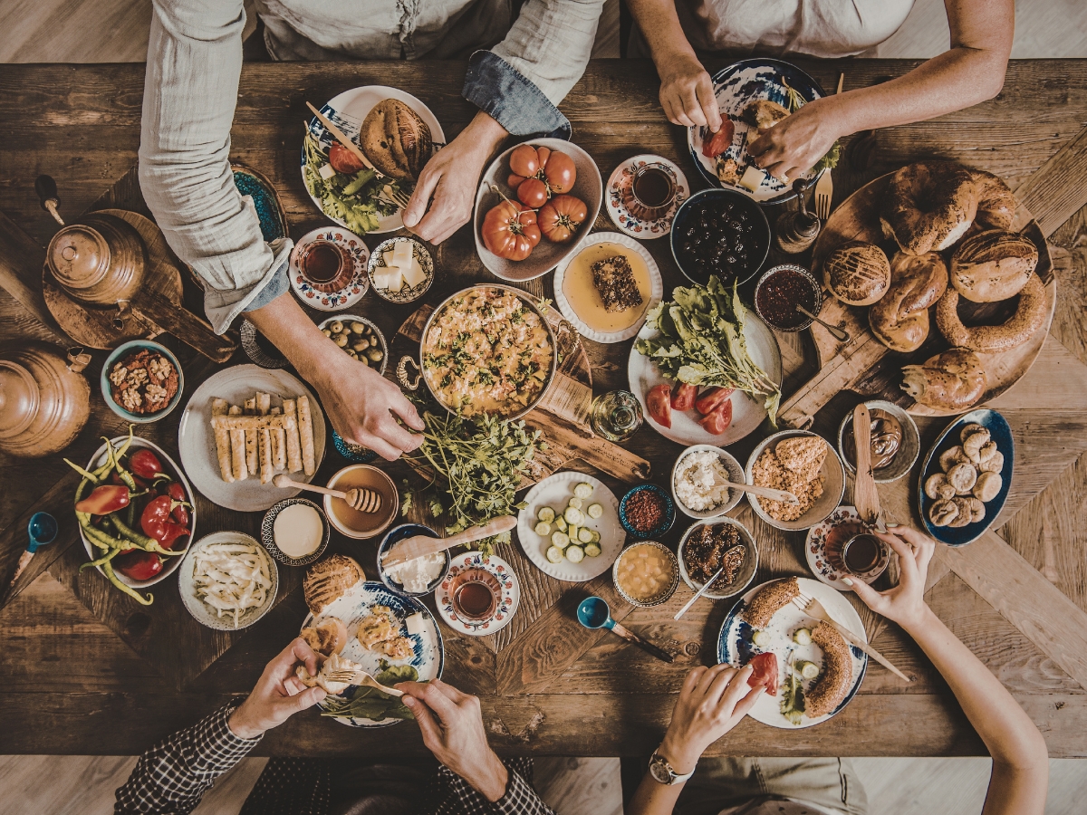 Overhead view of a lively dinner party with several people sharing a meal at a wooden table filled with various dishes, bread, vegetables, spreads, and drinks.