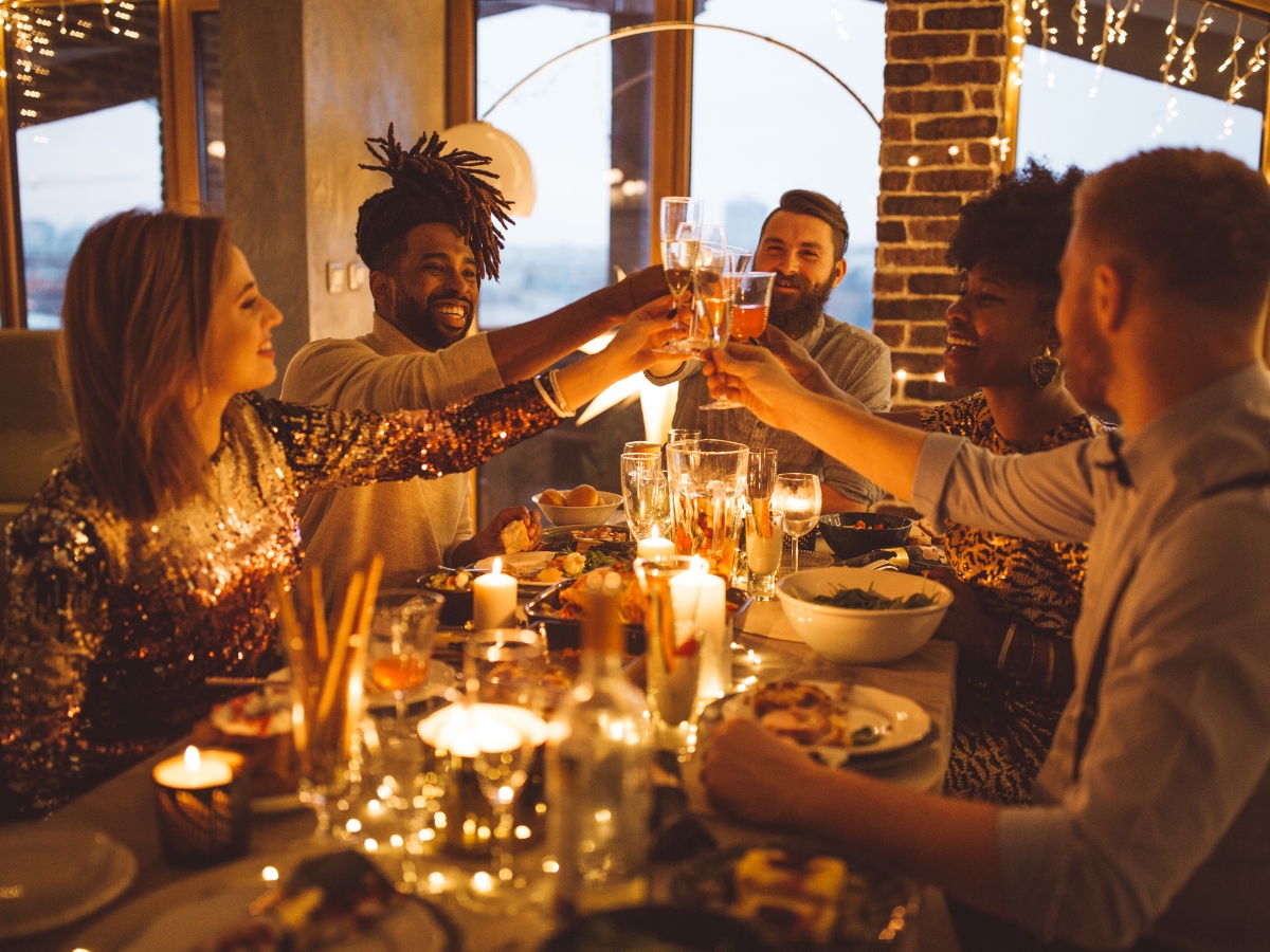 Five people sit around a decorated dinner table at a lively dinner party, raising glasses in a toast, with food, candles, and string lights creating a festive atmosphere.