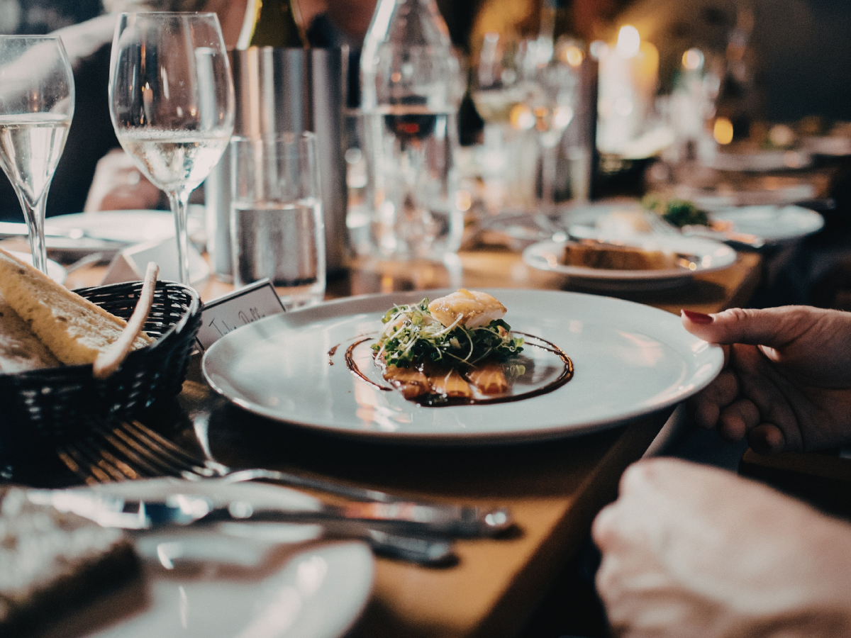 A person holds a plate with a gourmet dish at a dinner table set with wine glasses, water, bread, and utensils in a dimly lit restaurant, perfect for anniversary catering.