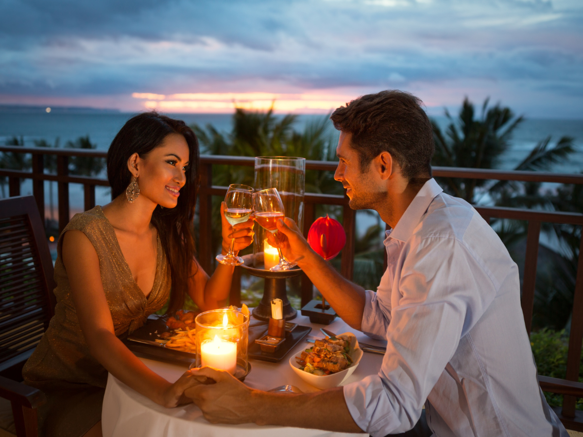 A couple sits at an outdoor table by candlelight, clinking wine glasses and enjoying anniversary catering with a sunset and ocean in the background.