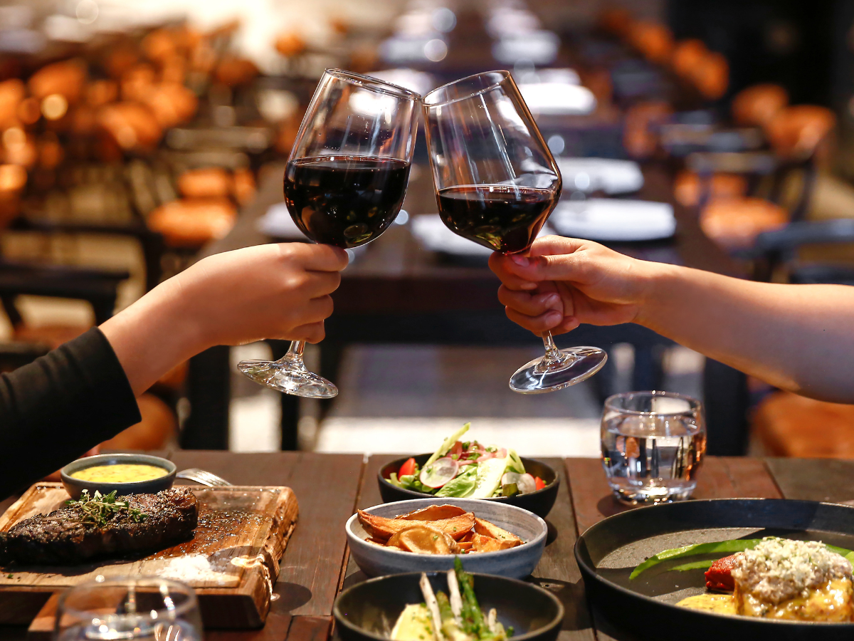 Two people clink glasses of red wine over a restaurant table set with steak, salad, fries, and water—perfect for a special night or intimate anniversary catering.