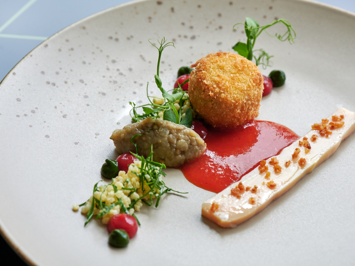 A plated gourmet dish perfect for anniversary catering, featuring a breaded, fried croquette, a sauce, a strip of terrine with garnish, diced vegetables, and artistic herb and puree accents on a white plate.