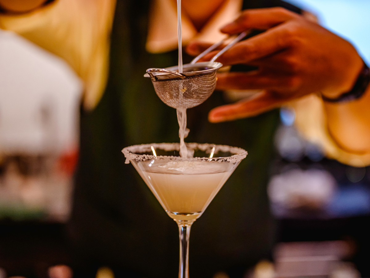 Cocktail Pairing Tips for Better Food and Drink Matches 1 A bartender pours a white cocktail through a metal strainer into a sugar-rimmed martini glass, ready for the perfect cocktail pairing.