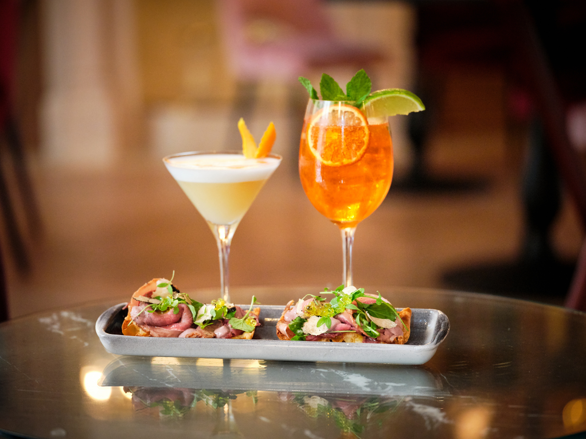 Cocktail Pairing Tips for Better Food and Drink Matches 2 Two cocktails, one creamy and one orange with lime and mint, are served as a perfect cocktail pairing on a glass table alongside three pieces of crostini topped with sliced meat and herbs.