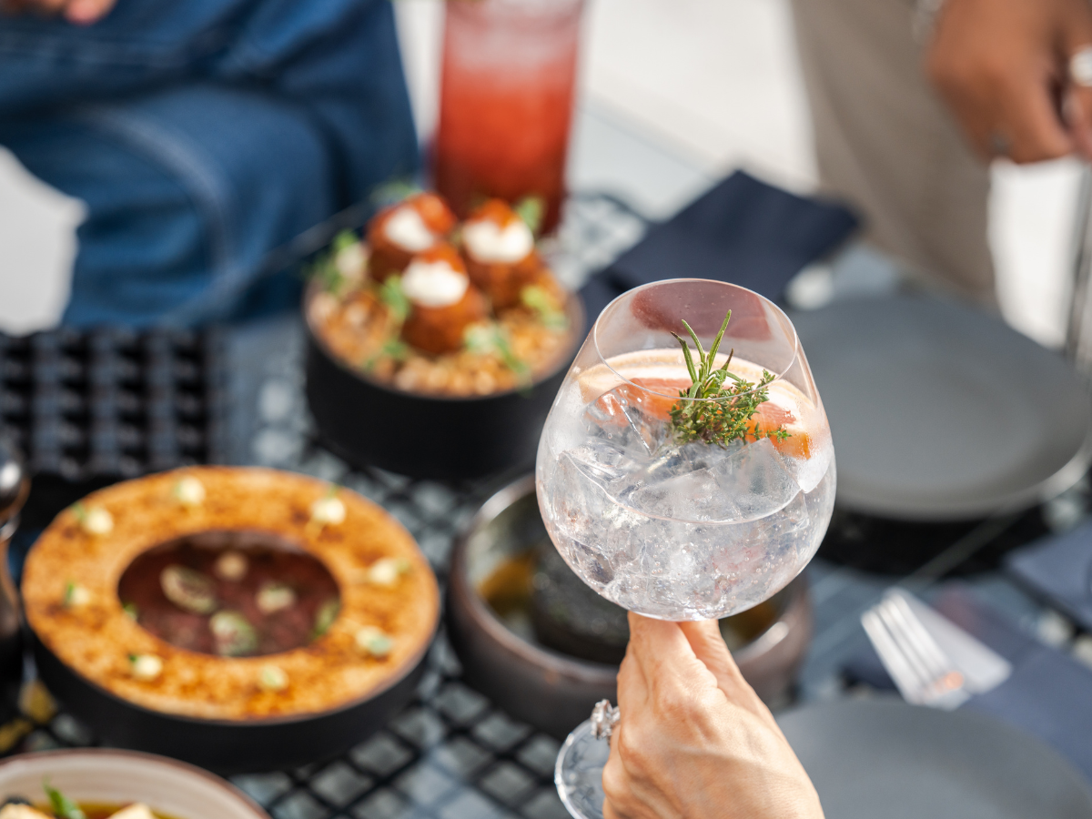 Cocktail Pairing Tips for Better Food and Drink Matches 3 A hand holds a gin and tonic with herbs and citrus garnish over a table set with various dishes and drinks, perfect for cocktail pairing.