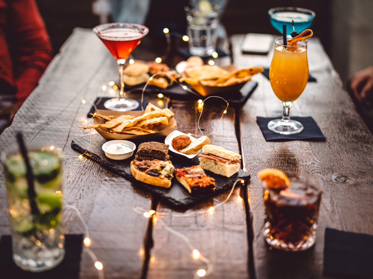 Cocktail Pairing Tips for Better Food and Drink Matches 4 A wooden table set with assorted cocktails, appetizers, breadsticks, and dipping sauces, decorated with string lights—perfect for a memorable cocktail pairing experience.