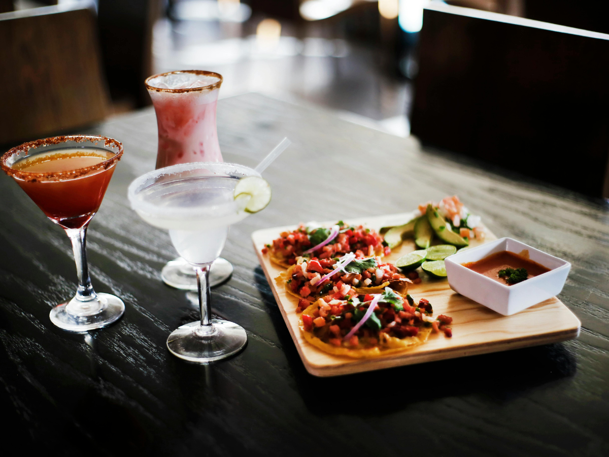 Three cocktails and a wooden platter with tostadas, lime wedges, and salsa create the perfect cocktail pairing experience on a dark table in a restaurant setting.
