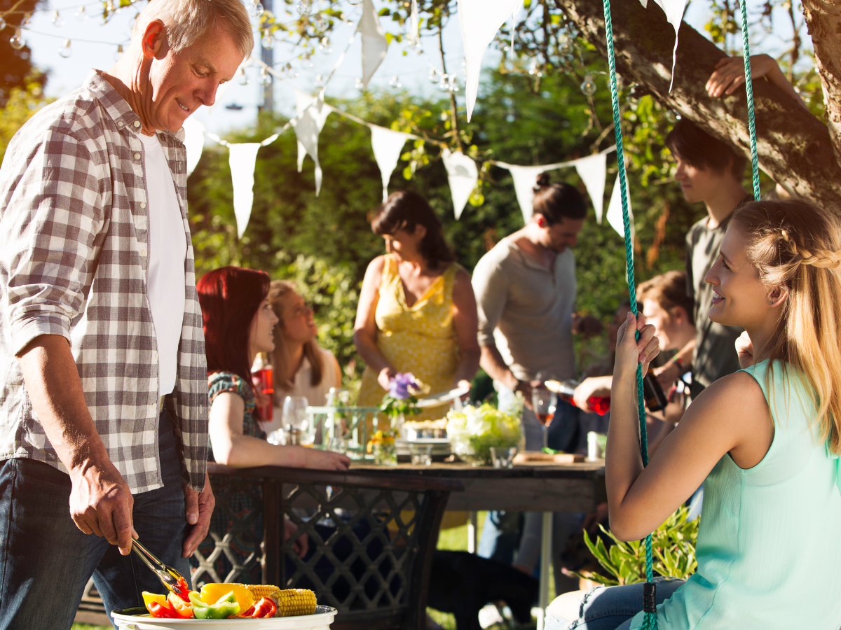 People gather outdoors for a summer BBQ picnic. A man grills vegetables while others sit or stand around a table decorated for a casual garden party.