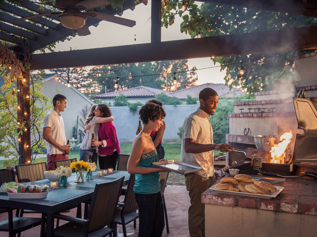 People gather on a patio at sunset for a summer BBQ picnic; one grills burgers, another holds a tray, while others chat or hug near the decorated outdoor dining table.