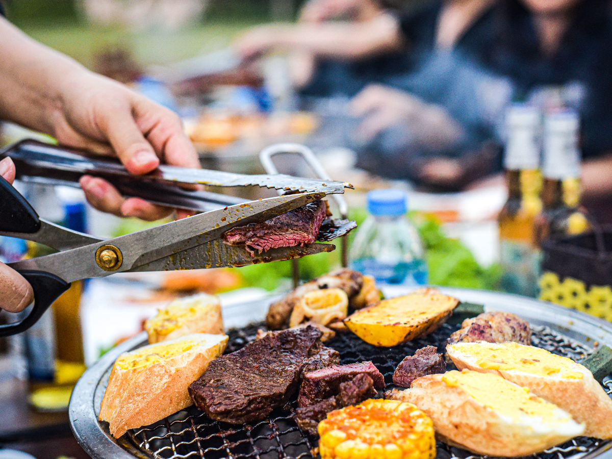 A person uses scissors to cut grilled meat over a barbecue filled with steak, corn, and bread at a lively summer BBQ picnic, with other people and food items in the background.