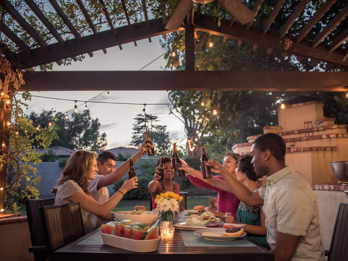 Six people sit around an outdoor table at dusk, raising drinks in a toast during a summer BBQ picnic. The scene is lit with string lights and decorated with flowers, while food like watermelon and burgers fills the festive table.