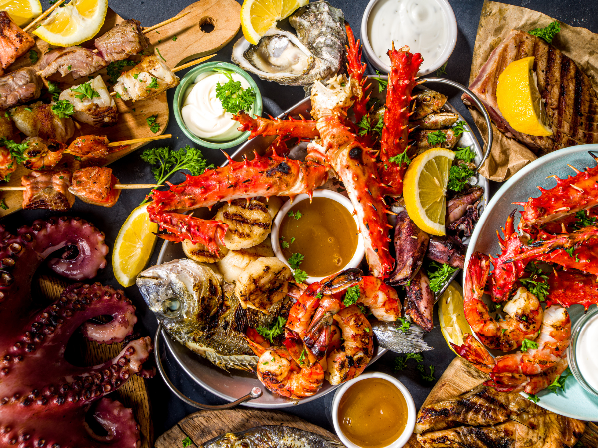 Assorted grilled seafood platter with crab legs, shrimp, octopus, oysters, fish, lemon wedges, and dipping sauces—perfect for a summer BBQ picnic. Skewers and fresh flavors await on the table.